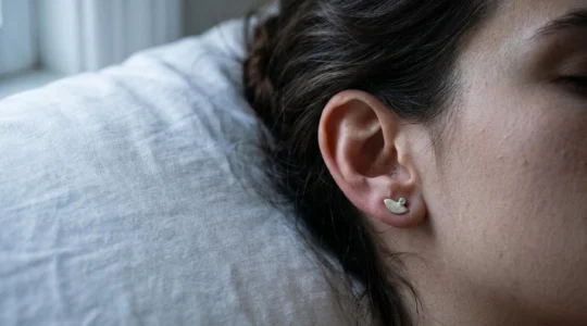 Gros plan sur une oreille féminine au repos sur un oreiller blanc immaculé la nuit, montrant une puce d'oreille discrète sans inconfort visible