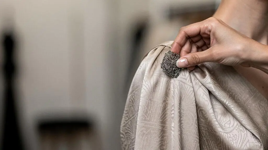Main féminine ajustant délicatement une broche ornée sur un tissu de soie fluide, illustrant une technique de fixation sans dommage